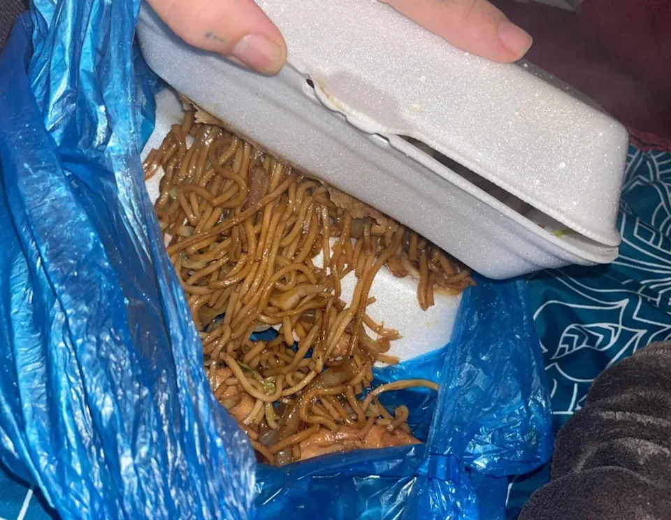 Takeout container with noodles, partially opened inside a blue plastic bag, on a patterned surface. A hand is opening the container