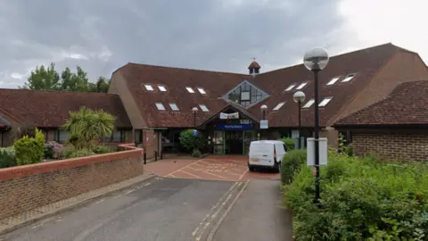 Google A brick building with a large sloped tile roof. There is a Blue sign over the entranceway that reads Priority House.