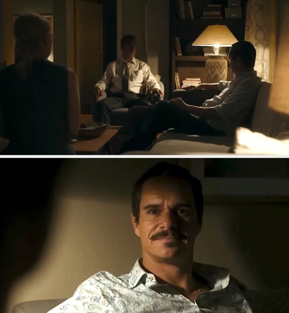 Scene from a TV show with three people in a dimly lit room. A man with a mustache is seated on a couch, engaging with the others
