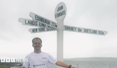 Drew Jonkmans holds a Guernsey flag above his head at the Land's End road sign. He is wearing a white t-shirt and blue shorts. A building is at the end of a road in background. The sea is behind Drew. It is a sunny day with a few clouds in the sky.