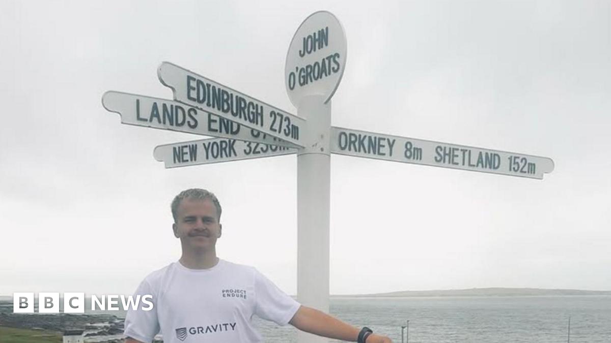 Drew Jonkmans holds a Guernsey flag above his head at the Land's End road sign. He is wearing a white t-shirt and blue shorts. A building is at the end of a road in background. The sea is behind Drew. It is a sunny day with a few clouds in the sky.