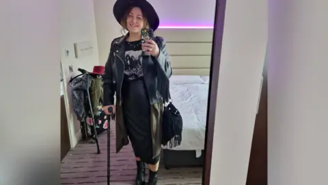 Stewarts Law Firm A woman in a black leather jacket, hat, dress and boots. She is standing with a walking stick. 