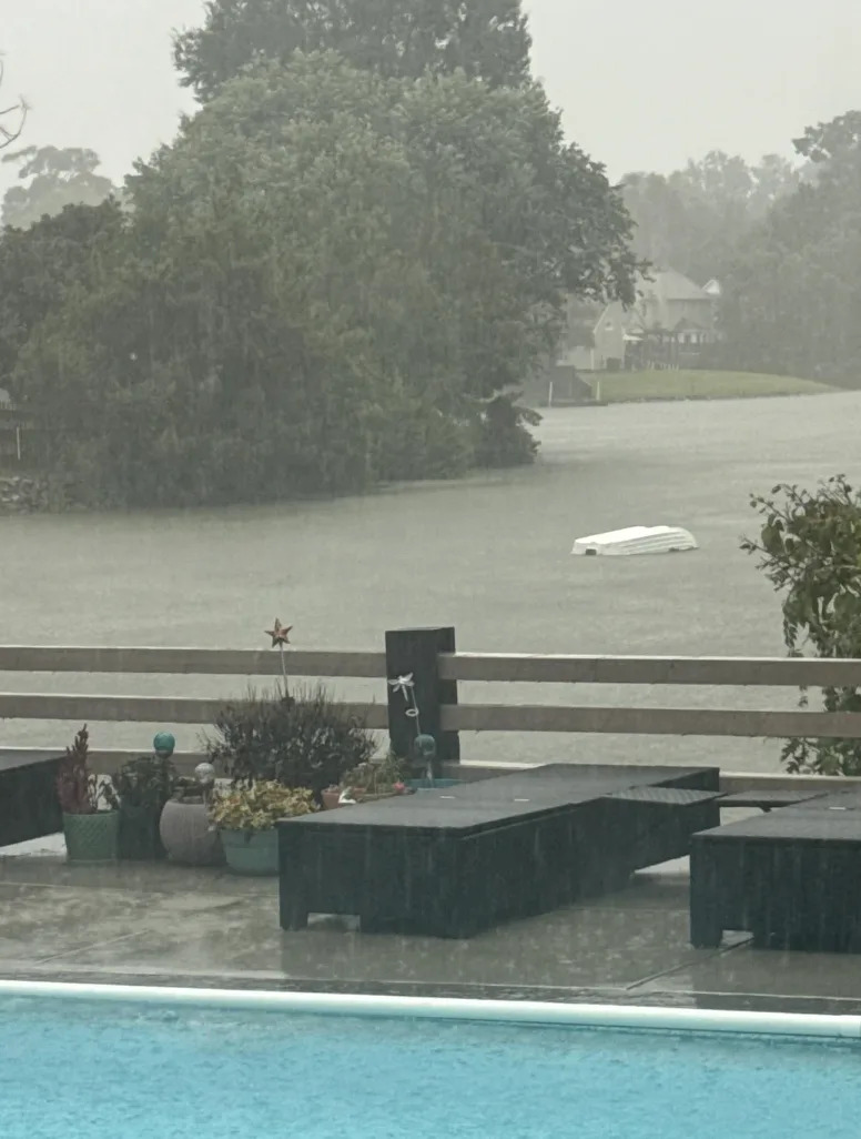 Rainy backyard scene with submerged boat on flooded lake, patio furniture in the foreground, and trees in the background