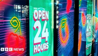 Kester and Zoe are sitting outside in a park looking directly at the camera. Kester is wearing a black tee shirt and thick, dark-rimmed glasses, and has a small tattoo on the left of his neck. Zoe has her brown hair tied in back and up in a bob and has a wry, closed-mouth, smile. She is wearing an orange, black and light grey baseball top.