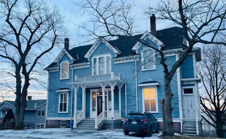 Light shines from one window of a two-story Victorian home with blue siding, white trim, and a steps leading to a a black double door. The trees out front are without their leaves and a car is parked in the driveway.
