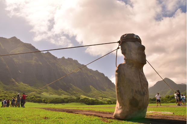 A moai replica is in the foreground and there are three ropes attached to the head with three teams pulling them
