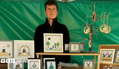 Aiden Thompson at is stall at Carlisle Teenage Market. His goods, including framed Christmas cards a pictures, are laid out on tables in front of him. He is wearing a black jumper and has dark hair and blue eyes.