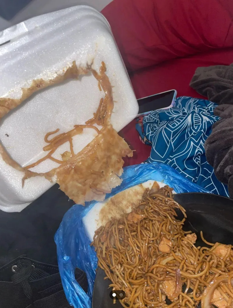 Takeout food spilled from a container onto a plastic bag, with noodles and sauce visible. Nearby are a phone and fabric with patterns
