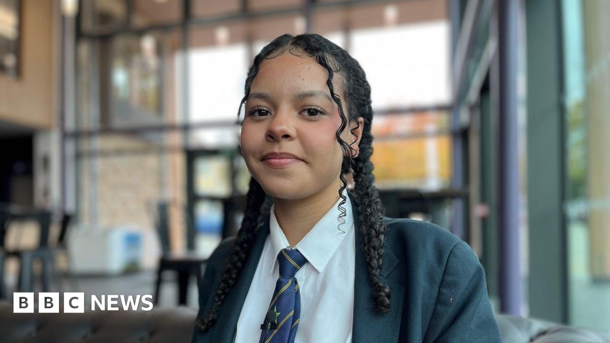Sam Coton wears a navy polo shirt with the 'Better Me, Better You' logo on it. He is sitting in the school theatre bar with floor to ceiling glass windows, you can see tables and chairs behind him. He is smiling at camera.