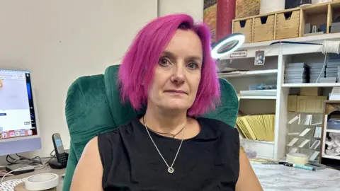 Claudia Sermbezis/BBC A woman with bright pink hair sat in a green chair and wearing a silver necklace. She is sat in front of shelves of mail materials like envelopes and a desk with a computer on it.