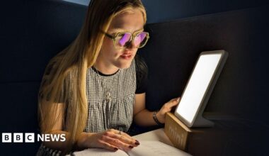 A young woman with long blond hair wearing glasses, a black T-shirt, and a black-and-white checkered dress. She is sitting at a desk or table and reading a book. Next to her is a bright, rectangular light therapy lamp. The light is illuminating her face and the book she is reading. Her nails are painted red with white polka dots.