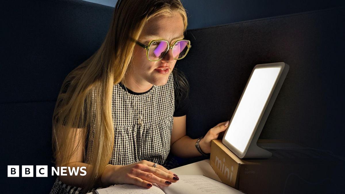 A young woman with long blond hair wearing glasses, a black T-shirt, and a black-and-white checkered dress. She is sitting at a desk or table and reading a book. Next to her is a bright, rectangular light therapy lamp. The light is illuminating her face and the book she is reading. Her nails are painted red with white polka dots.