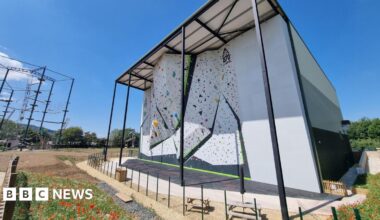 In bright sunshine and near cloudless skies, a giant climbing wall towers over the surrounding area. Several overhangs protrude out from the main structure.