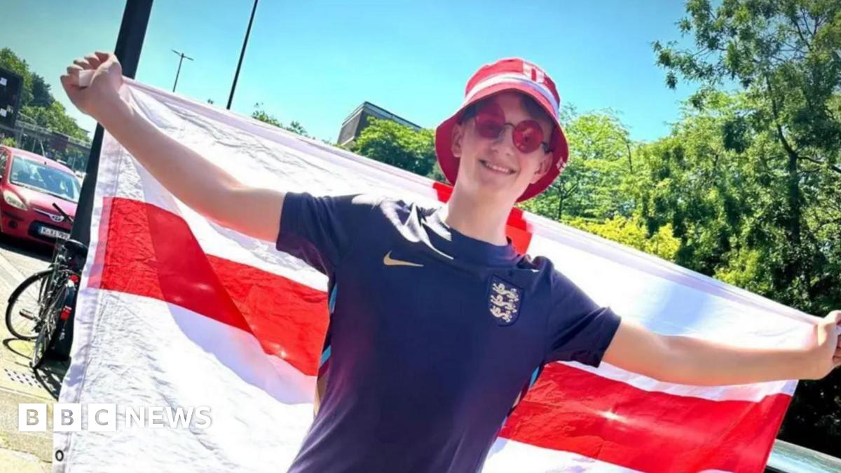 A young man wearing sunglasses, a blue football shirt and a bucket hat with his arms outstretched, hold an England flag behind him
