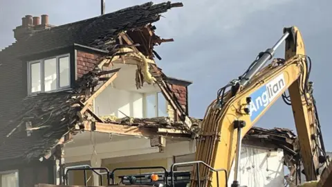 Richard Daniel/BBC A large yellow crane in front of a partially demolished home. It is cloudy above.