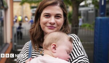 A portrait of Holly and her young daughter Ettie. Holly is a young woman aged 30 with long brown hair and  wearing a striped blue top. She is carrying her baby Elly who is asleep in her arms and wearing a very light pink top with long sleeves.