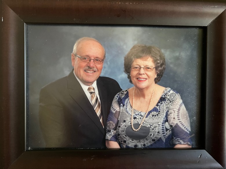 A white senior couple, the balding smiling man in a black jacket, white shirt and tie, the smiling woman in a blue dress, styled brown hair, glasses, and pearl earrings and necklace, are seen in a portrait held in a dark brown frame.