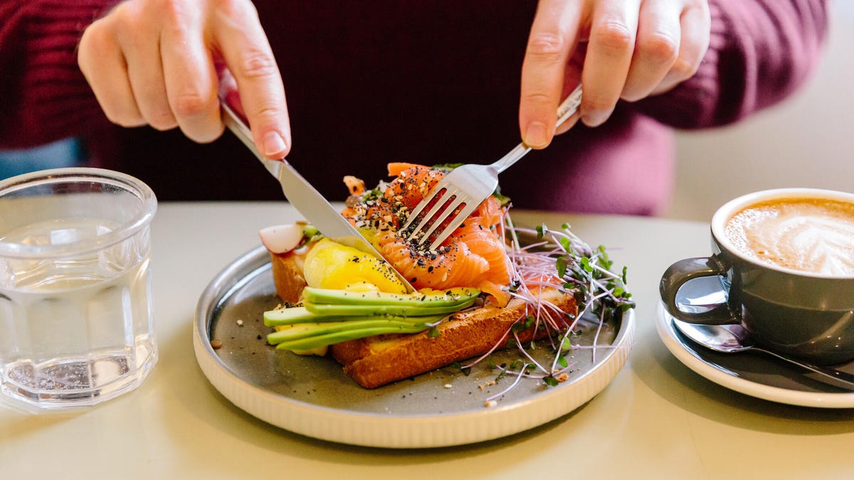 A man's hands cutting a salmon and avocado flatbread with a knife and fork.