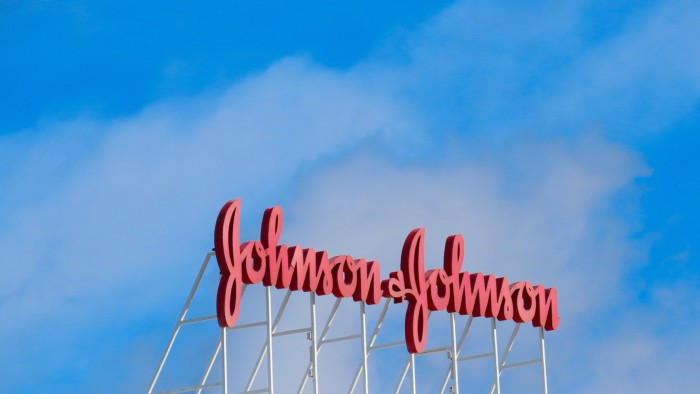 Red Johnson & Johnson logo in script font mounted on a rooftop against a blue sky with scattered clouds.