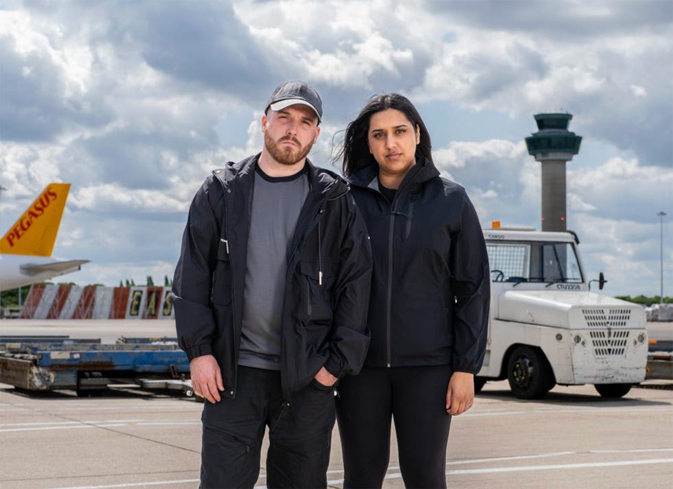 two individuals standing on an airport tarmac with an air traffic control tower in the background and a cargo vehicle nearby