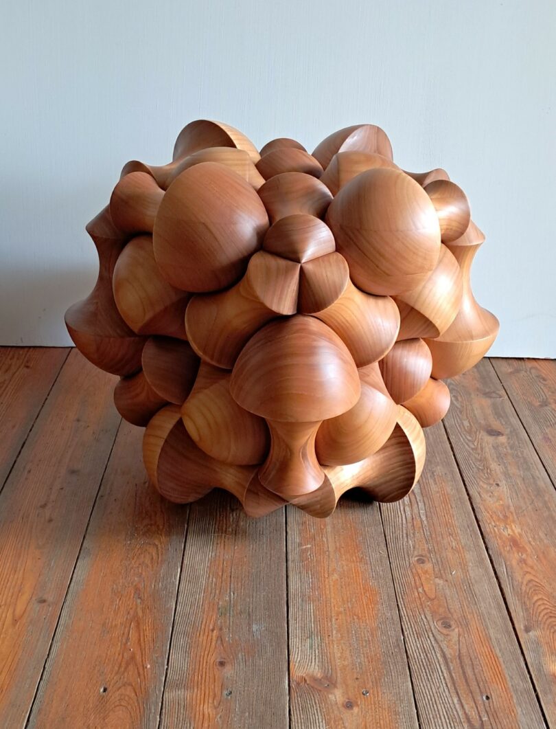 A wooden sculpture with an intricate geometric design featuring rounded, bulbous shapes, placed on a wooden floor against a plain white wall