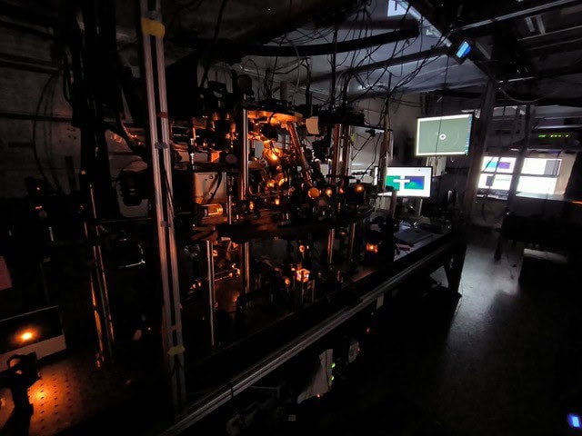 Photo of a darkened optics laboratory with screens and a vacuum system. The scene is bathed in orange light from the lasers used