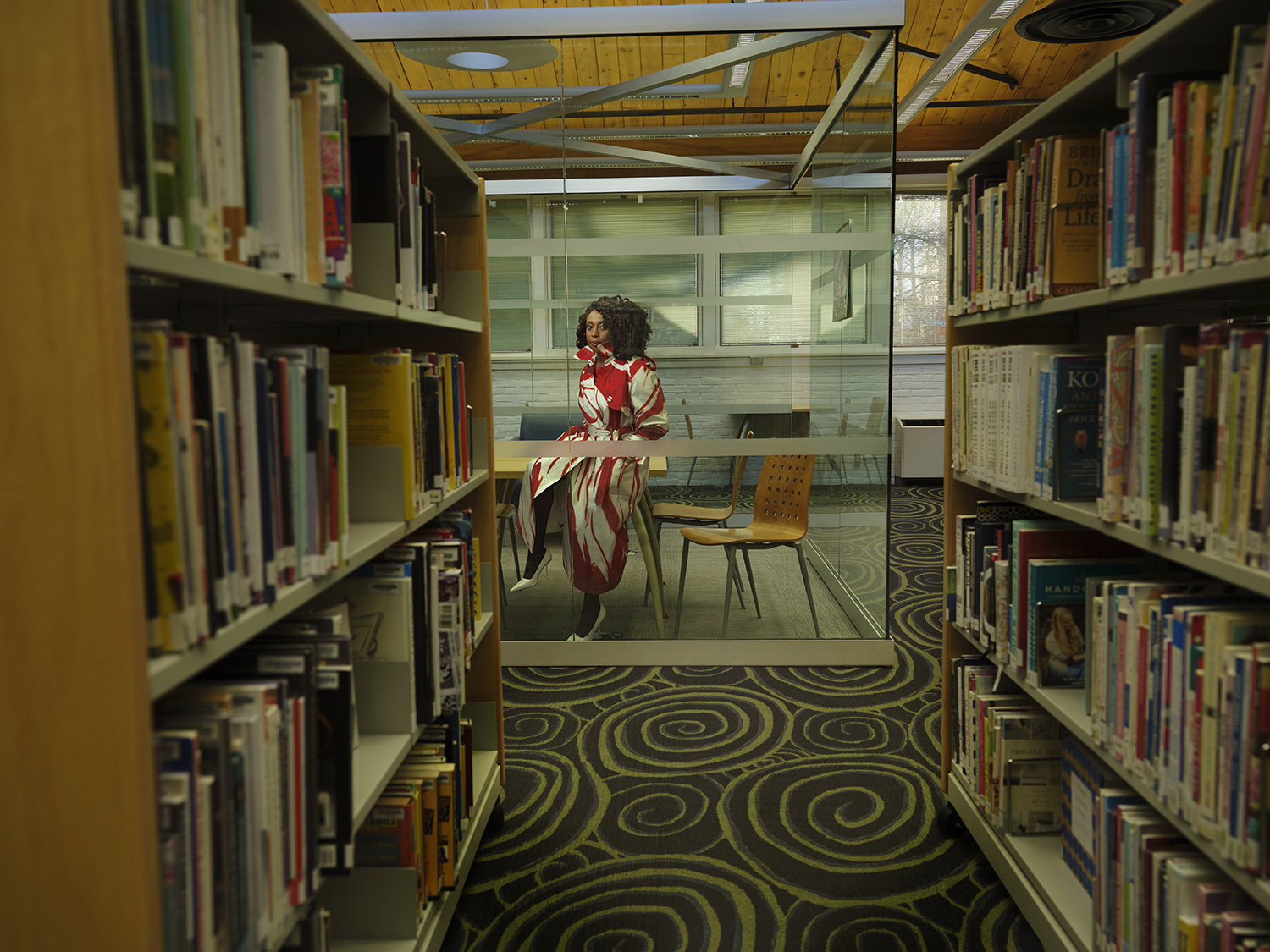 Chimamanda Ngozi Adichie sitting in a glass-enclosed room in a library.