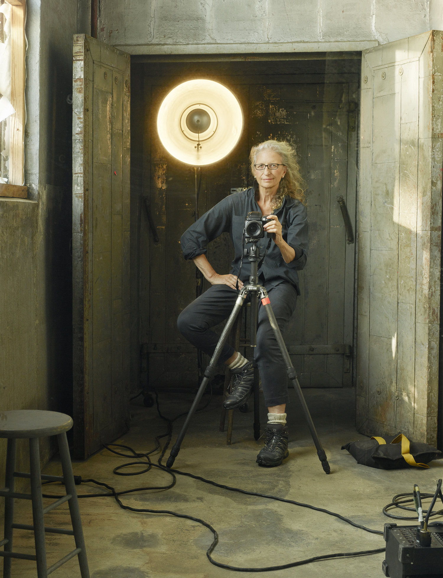 Annie Leibovitz holding a camera, sitting on a stool in a studio.