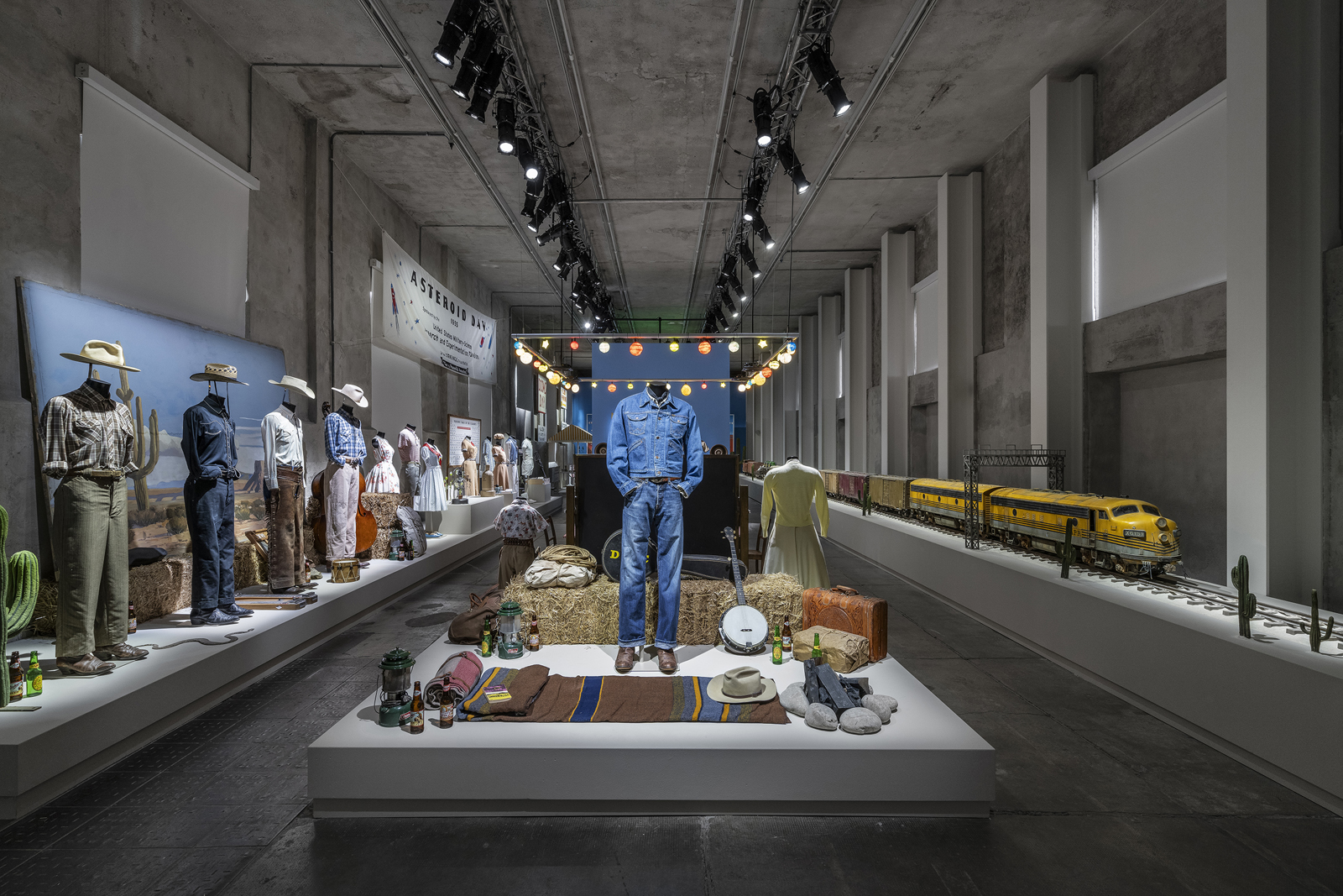 Exhibition with mannequins dressed in western wear, a denim outfit on a central platform, and a model train set.