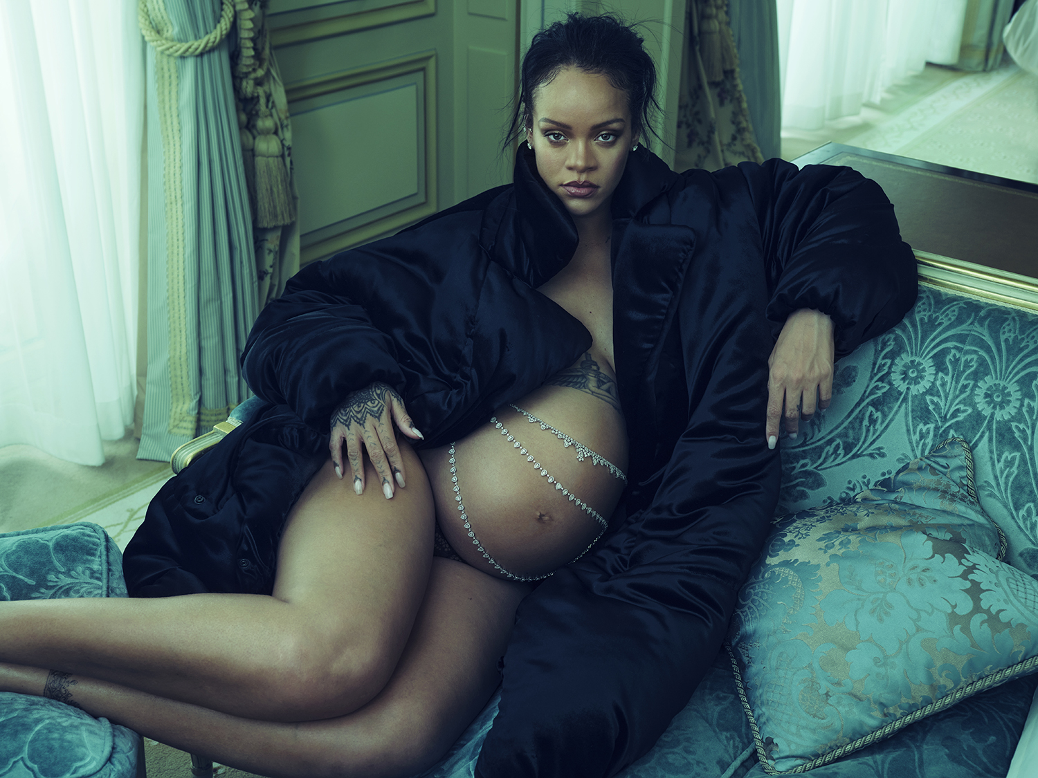 Rihanna pregnant, wearing a black puffer coat and diamond body chains, reclining on a velvet couch.