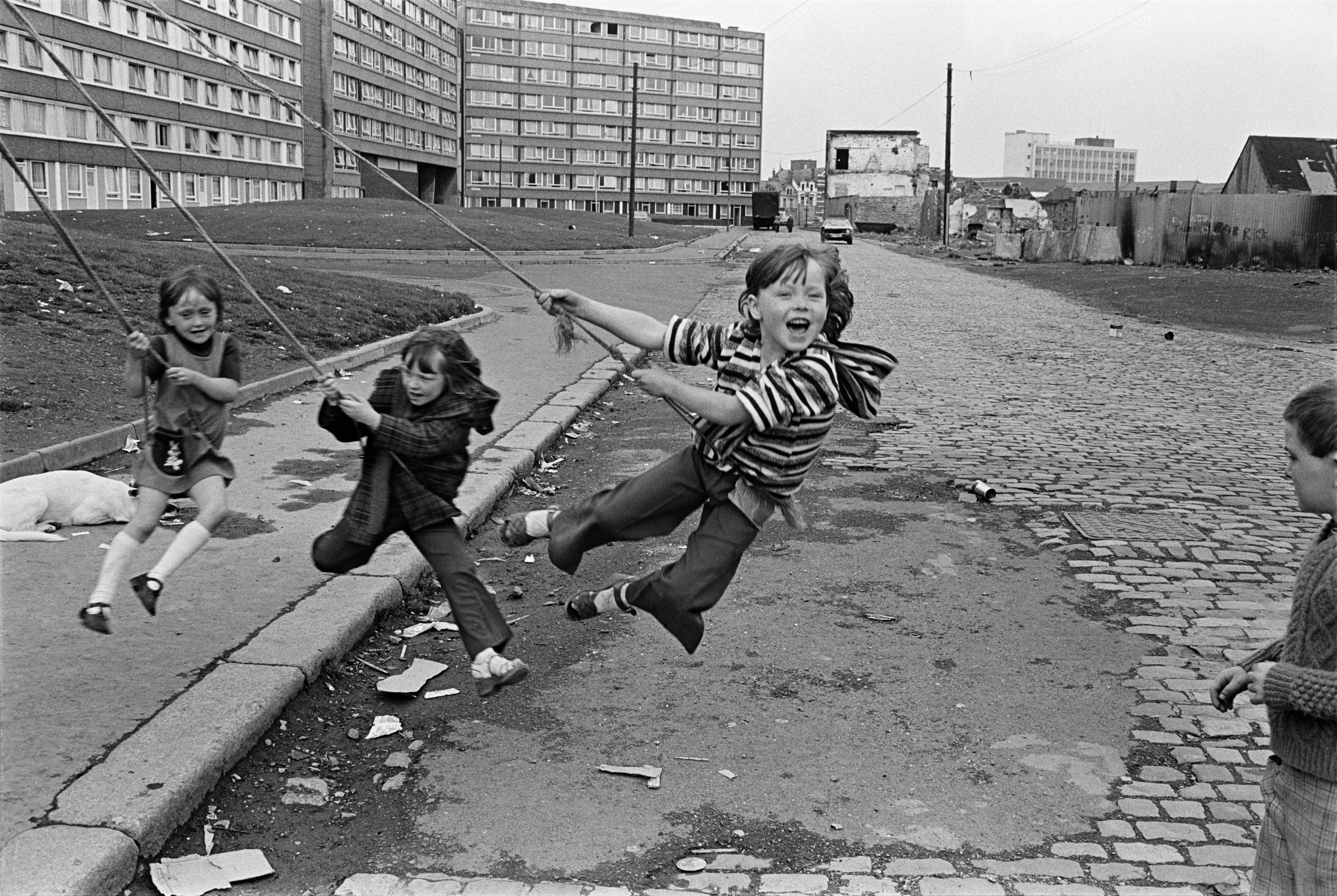 Three children playing on rope swings outside Divis Flats in West Belfast.