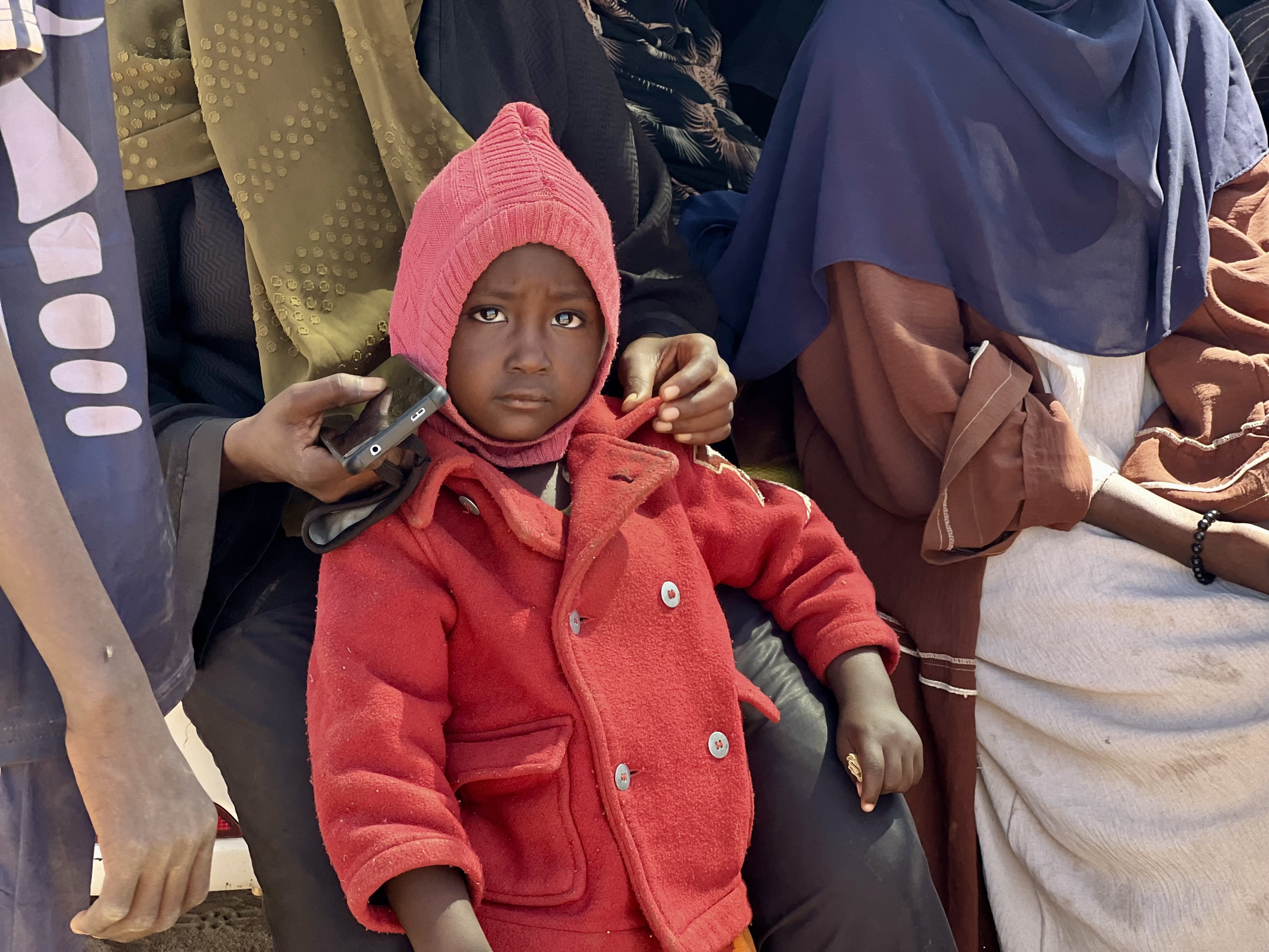 A displaced Sudanese child wearing a red coat and hat sits among adults.