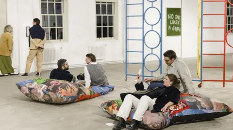 Trackie McLeod People are pictured sitting on beanbags chatting in the middle of the exhibition space. The climbing frame is pictured in the background, as well as people viewing various artworks around the room. 