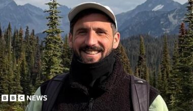 Oliver Gooch standing smiling in front of trees and mountains smiling. He has dark hair and a beard and is wearing a white baseball cap and black fleece and scarf