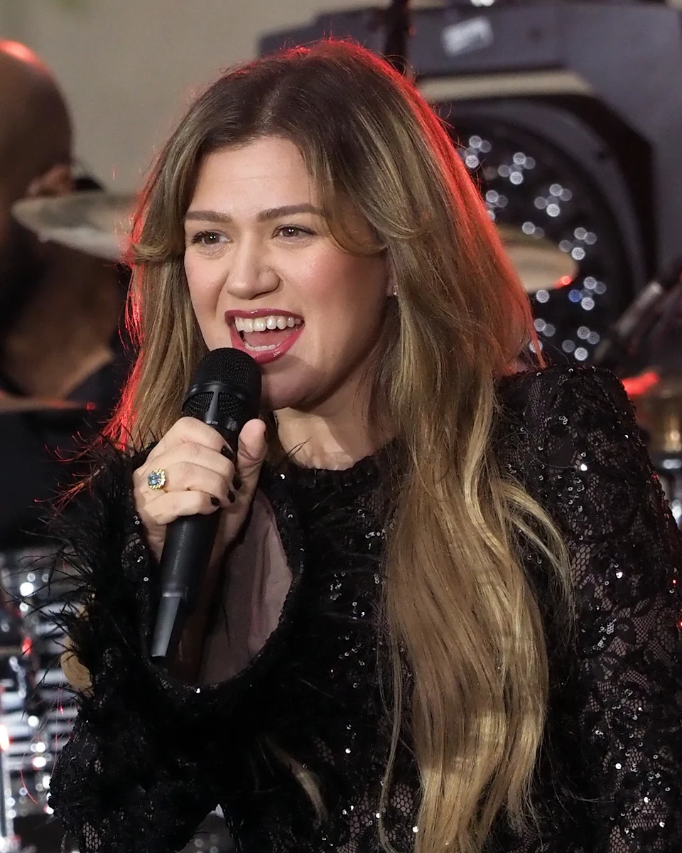 A performer singing into a microphone, wearing a glamorous black sequined top, with long wavy hair