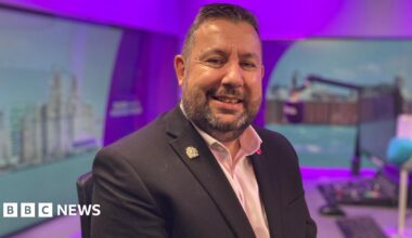 Anthony Burns sits in a radio studio, a blue microphone and control desk are visible in the background. He smiles at the camera. He has short dark hair and a stubbly beard. He wears a dark suit jacket with a pink, open-collar shirt underneath.