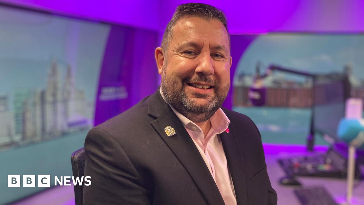 Anthony Burns sits in a radio studio, a blue microphone and control desk are visible in the background. He smiles at the camera. He has short dark hair and a stubbly beard. He wears a dark suit jacket with a pink, open-collar shirt underneath.