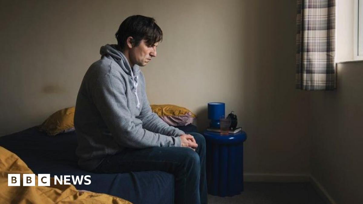 A wearing a grey hood and jeans is sitting on a bed.  He has a sad look on his face and he is looking down at the floor, with his hands clenched.