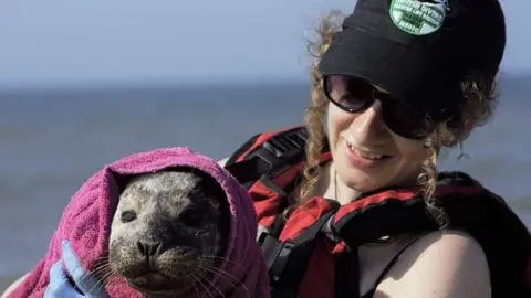 Gem Simmons A woman wearing sunglasses looks and smiles at a seal that is wrapped in a purple blanket 