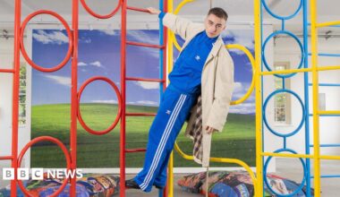 Trackie is pictured in a blue tracksuit, hanging from a colourful climbing frame in his exhibition. In the background, there is more of his artwork pictured, like a big windows home screen display, and beanbags with wrestling images on them.