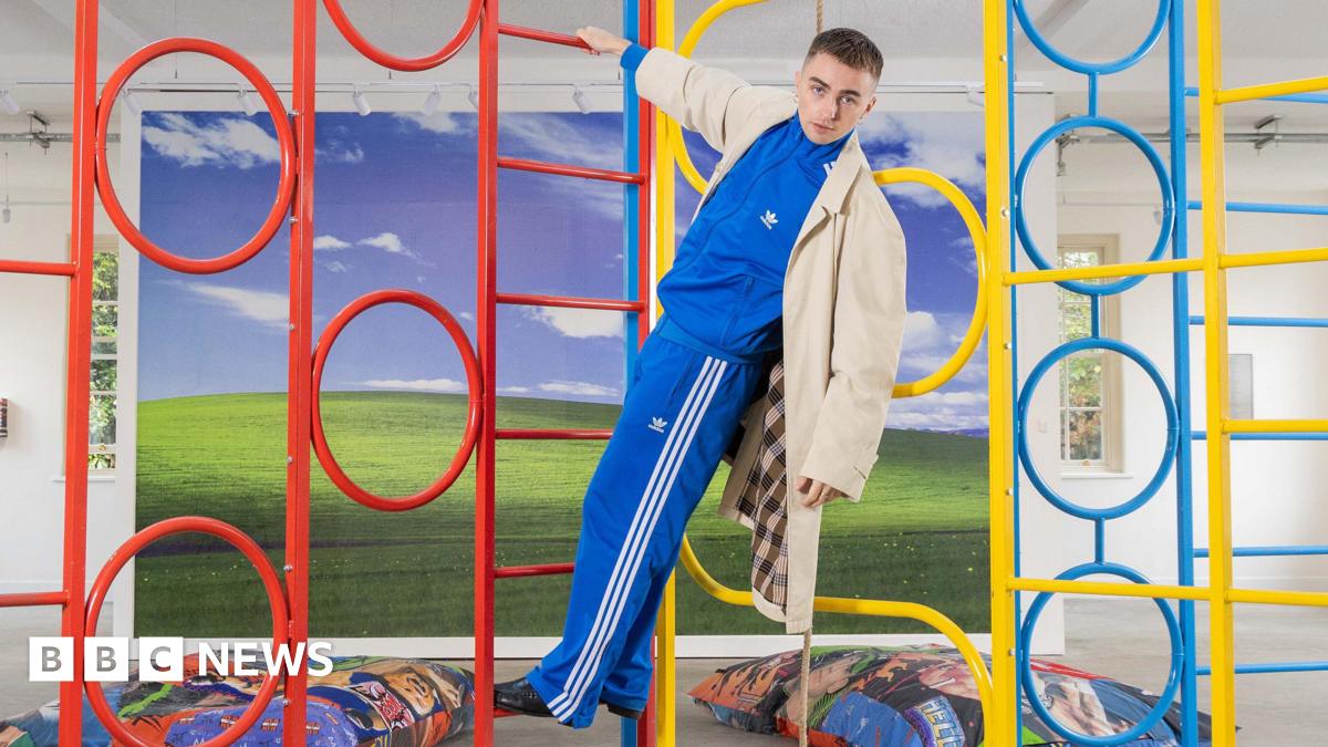 Trackie is pictured in a blue tracksuit, hanging from a colourful climbing frame in his exhibition. In the background, there is more of his artwork pictured, like a big windows home screen display, and beanbags with wrestling images on them.