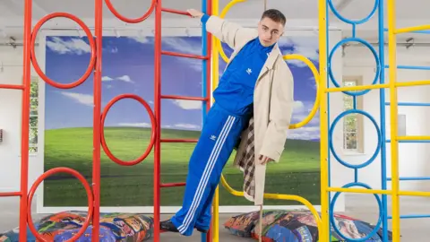 Trackie McLeod Trackie is pictured in a blue tracksuit, hanging from a colourful climbing frame in his exhibition. In the background, there is more of his artwork pictured, like a big windows home screen display, and beanbags with wrestling images on them. 