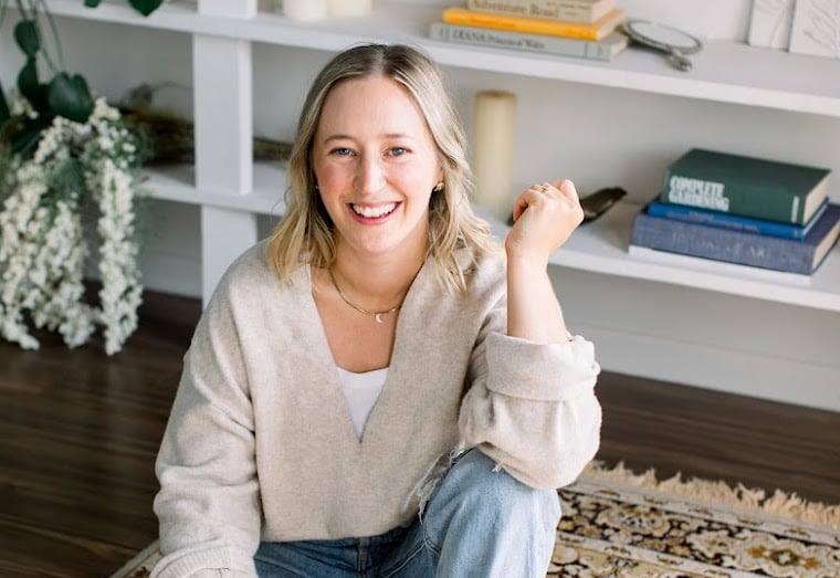 Woman with blonde hair smiling, sitting cross-legged on a patterned rug in a bright room with built-in shelving