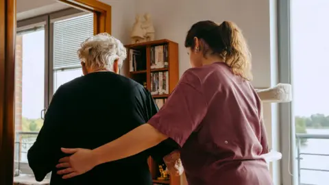 Getty Images An elderly woman with short white hair is escorted by a woman with long brown, wearing maroon scrubs. The younger of the two has her left hand on the woman's back. The elderly woman wears a black dress. A wooden book shelf, two shelves wide and four long is partially stacked with books. There are two windows either side of the bookshelf, a body of water is visible outside. 
