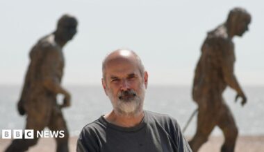 Laurence Edwards in a grey T-shirt standing in front of two statues on a beach. He has grey hair and a thick grey beard. The statues are bronze and look as if they are walking. The North Sea is in the background.