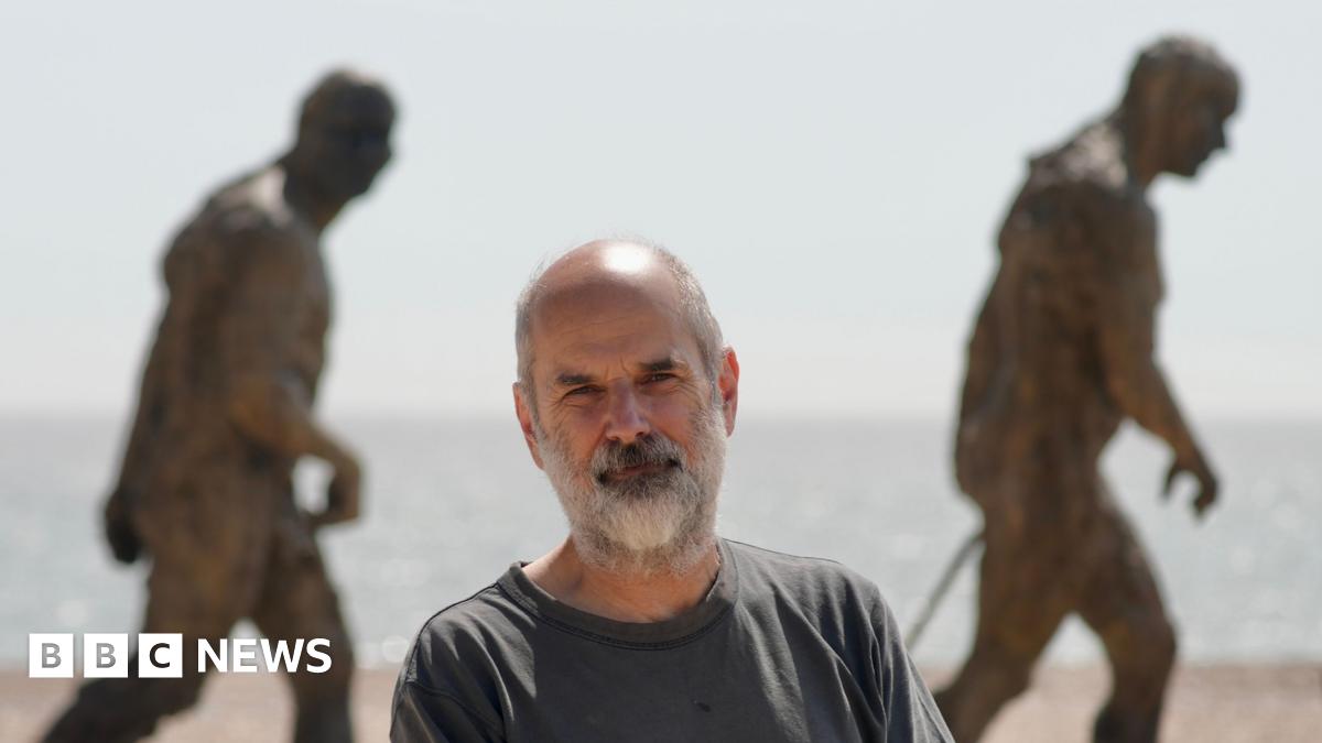 Laurence Edwards in a grey T-shirt standing in front of two statues on a beach. He has grey hair and a thick grey beard. The statues are bronze and look as if they are walking. The North Sea is in the background.