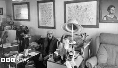 Black & white photo with Henry Orlik in his 70s in a living room with framed paintings behind him as she sits down, with a sofa to the right
