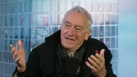BBC Robert De Niro, sitting in front of a blue background, wears a black coat and a dark scarf. He has short grey hair and is gesturing with both hands out in front of him. 