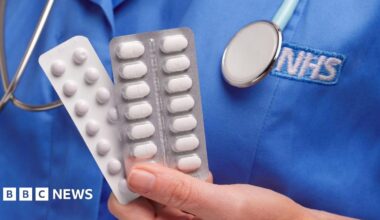 Close up of a hand holding two packets of white tablets. A blue nurses top is in the background with the letters NHS written on in white