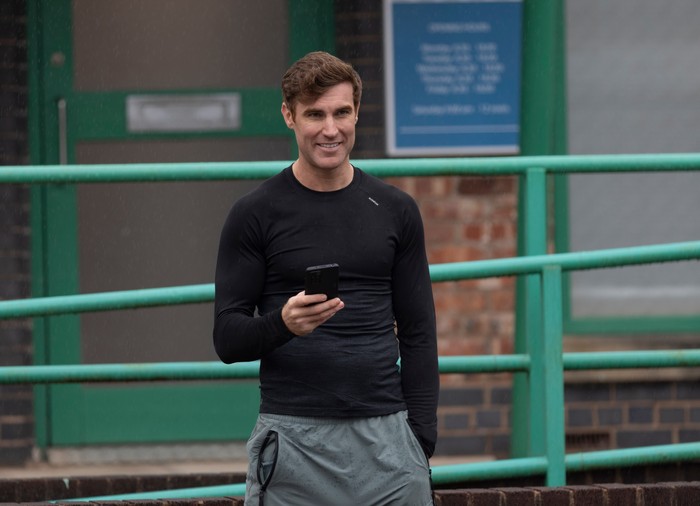 Theo Silverton smiles with his phone in his hand in Coronation Street
